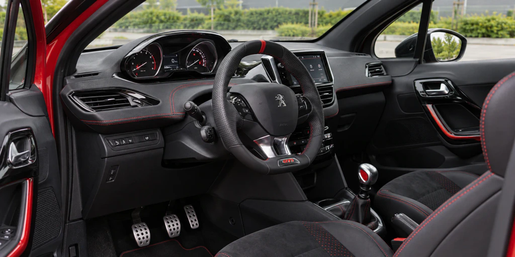 Illustrative left-hand-drive Peugeot 208 GTi cockpit with a small steering wheel, raised instrument cluster, manual gear lever and front seats.